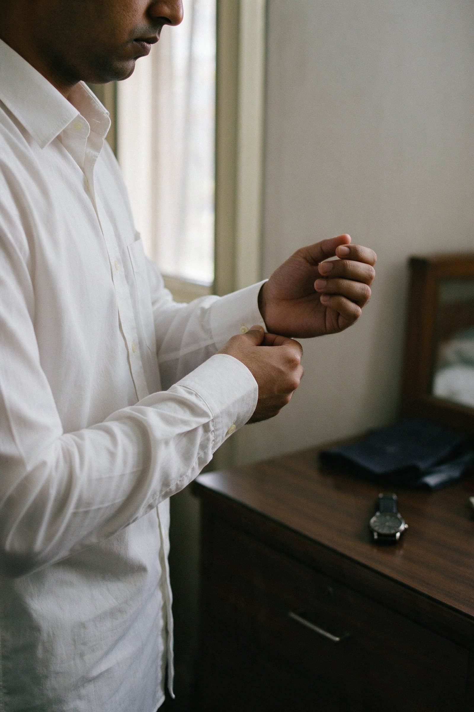 A man in a white shirt buttoning his cuff by a window, his watch waiting on the dresser