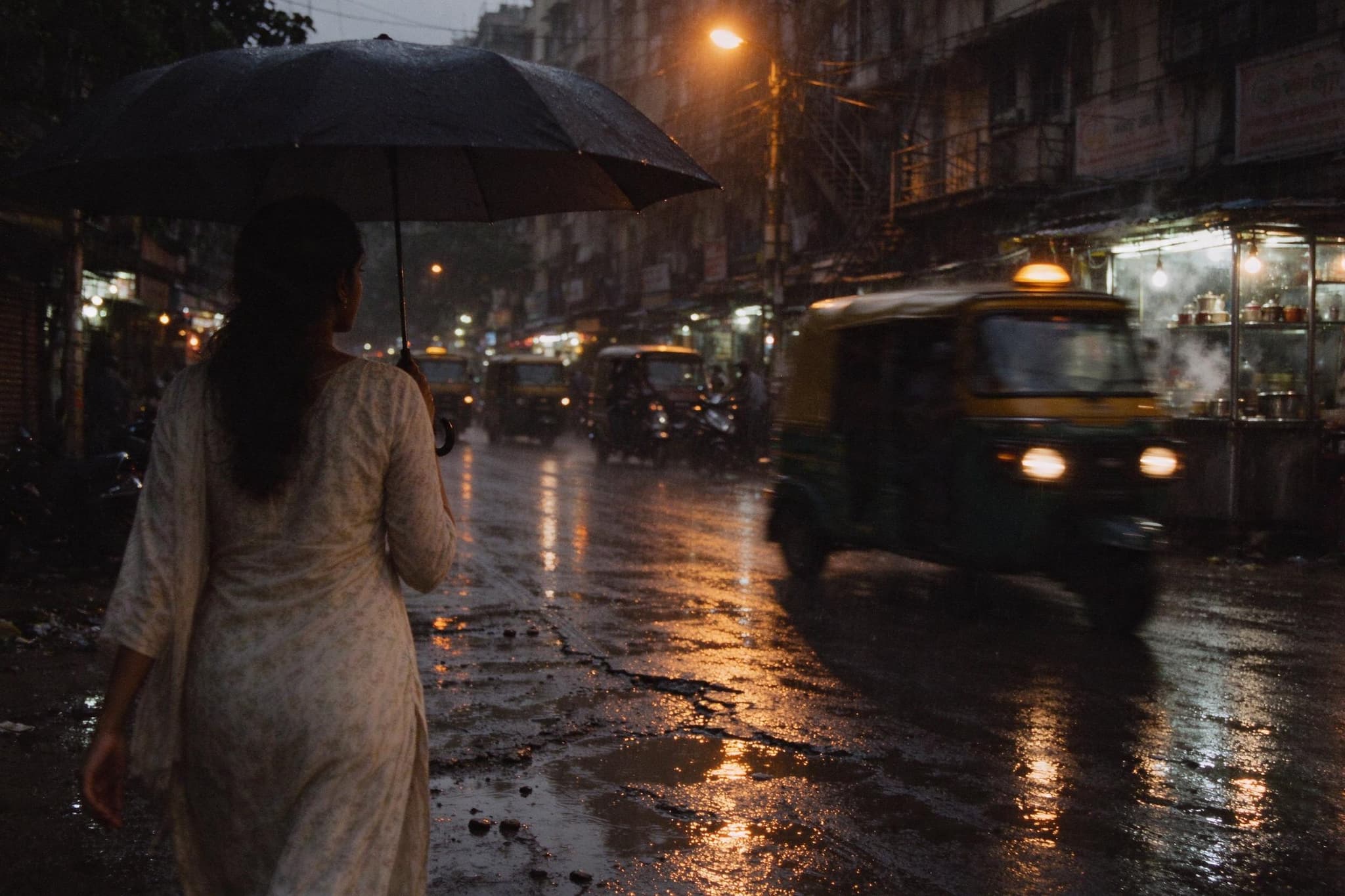 A woman in a pale kurta walking under an umbrella down a rainy Mumbai street at dusk, an auto-rickshaw passing